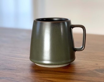 Può includere: Una tazza in ceramica verde scuro con fondo arrotondato e parte superiore leggermente svasata. La tazza ha una finitura liscia e lucida e un manico sul lato destro.