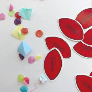 May include: Red American football-shaped stickers with white borders and black stitching are scattered on a white surface.  Colourful confetti in various shapes and hues, including pink, yellow, and blue, adds a playful touch to the composition.