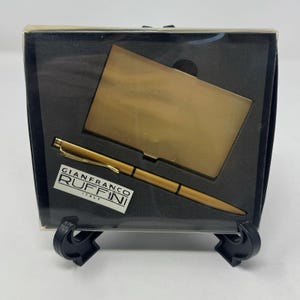 Könnte beinhalten: Ein goldfarbenes Stift- und Visitenkartenhalter-Set von Gianfranco Ruffini Italy, präsentiert in einer schwarzen Box. Der Stift ist schlank mit einem goldenen Clip, und der Kartenhalter ist rechteckig. Das Set wird auf einem schwarzen Ständer präsentiert.