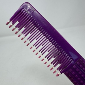 May include: A purple comb with silver and pink accents on the teeth. The comb is angled, showing the details of the teeth and the overall design. The comb is a beauty tool.