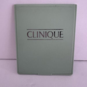 Puede incluir: Un estuche compacto con una tapa verde claro y la palabra "CLINIQUE" impresa en letras plateadas.