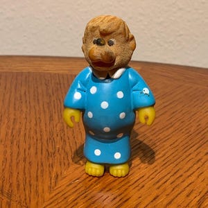 May include: A vintage figurine of a monkey wearing a blue polka-dot nightgown. The monkey has a brown head, yellow hands and feet, and a white collar. The figurine is standing on a wooden surface.