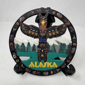 Puede incluir: Una pieza decorativa con temática de Alaska con un diseño de tótem. El tótem es marrón con detalles en negro, azul y amarillo, sobre un fondo de montañas y árboles. La palabra "ALASKA" se muestra en amarillo.