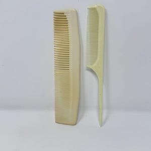 Könnte beinhalten: Zwei cremefarbene Vintage-Kämme aus Kunststoff. Einer ist ein Standardkamm mit eng stehenden Zähnen. Der andere ist ein feinzahniger Kamm mit spitzem Griff, der oft zum Stylen verwendet wird. Beide Kämme befinden sich vor einem weißen Hintergrund.