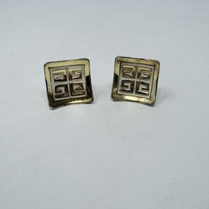 May include: A pair of square, gold-toned earrings. Each earring features a geometric design within a square frame. The design is composed of repeating shapes, creating a symmetrical pattern. The earrings are set against a white background.