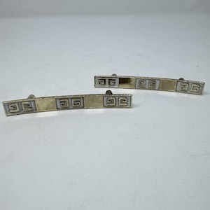 以下が含まれることがあります： 2つのヴィンテージゴールドトーンメタルネクタイクリップ。各クリップは、中央のゴールドトーンパネルと白と金の幾何学模様のアクセントを備えた長方形のデザインが特徴です。クリップは白い表面に表示されています。