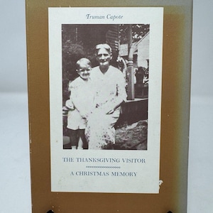 May include: A rectangular book with a tan cover featuring a central white panel. The panel displays a black and white photograph of a child and an adult, with the text Truman Capote, THE THANKSGIVING VISITOR, and A CHRISTMAS MEMORY printed in black.