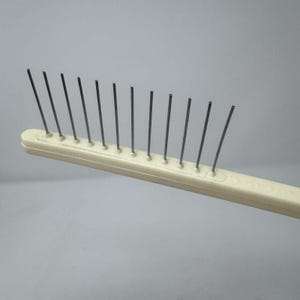 May include: A cream-colored pet grooming comb with a row of thin, metal tines. The comb's handle is rectangular and slightly curved, with the tines evenly spaced along one side. The tines are angled slightly away from the handle.