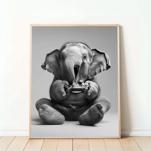 Puede incluir: Impresión enmarcada en blanco y negro que muestra a un elefante bebé jugando a un videojuego. El elefante está sentado, sosteniendo un mando de juego con su trompa. La impresión está en un marco de madera clara, sobre un fondo gris neutro.