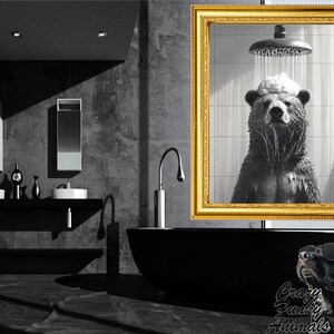 Op de afbeelding: Een zwart-witfoto van een beer die doucht. De beer staat in een douchecabine met water dat over zijn hoofd stroomt. De foto is ingelijst in een gouden lijst en hangt aan een muur in een badkamer.