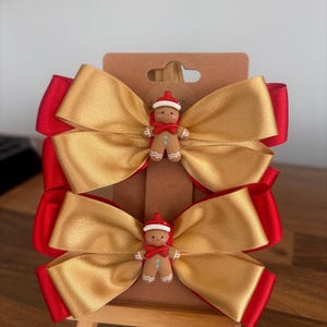 May include: Two layered hair bows featuring a metallic gold satin top layer and a solid red satin bottom layer. Each bow is centred with a small, smiling gingerbread person figurine wearing a red hat and scarf, attached to a brown display card.