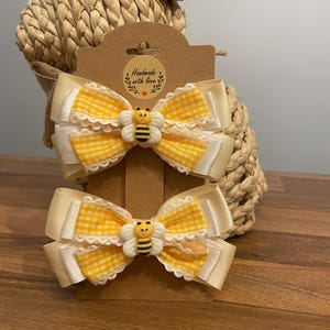 May include: Two hair bows with yellow gingham and cream-coloured ribbons, each adorned with a small bee embellishment. The bows are attached to a brown card with the text "Handmade with love". A woven basket is in the background.