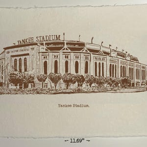 Op de afbeelding: Een sepia-getinte prent van Yankee Stadium, met een afmeting van 21 x 29,7 cm. Het stadion is afgebeeld in een gedetailleerde architectonische tekenstijl, met de woorden "Yankee Stadium" boven de structuur. De prent is op getextureerd papier.