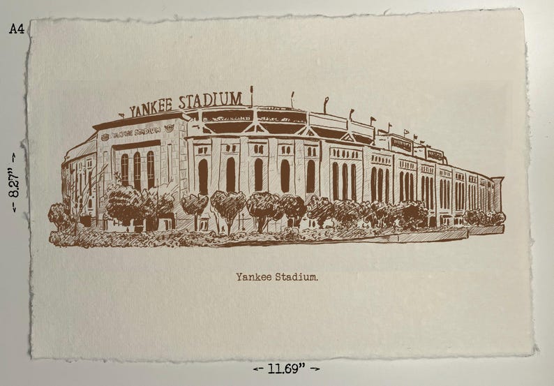 Op de afbeelding: Een sepia-getinte prent van Yankee Stadium, met een afmeting van 21 x 29,7 cm. Het stadion is afgebeeld in een gedetailleerde architectonische tekenstijl, met de woorden "Yankee Stadium" boven de structuur. De prent is op getextureerd papier.