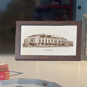Op de afbeelding: Een ingelijste prent van Yankee Stadium in sepiatinten. Het kunstwerk toont de buitenkant van het stadion met de tekst "YANKEE STADIUM" erboven en "Yankee Stadium" eronder. De lijst is donkerbruin.