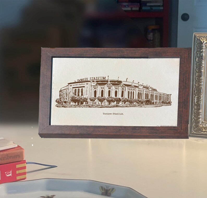 Op de afbeelding: Een ingelijste prent van Yankee Stadium in sepiatinten. Het kunstwerk toont de buitenkant van het stadion met de tekst "YANKEE STADIUM" erboven en "Yankee Stadium" eronder. De lijst is donkerbruin.