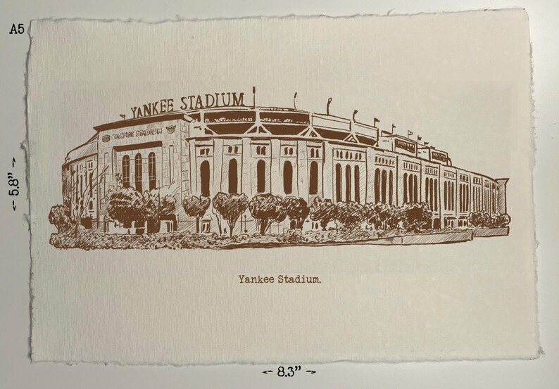 Op de afbeelding: Een bruine inkttekening van Yankee Stadium op getextureerd, gebroken wit papier. Het stadion is gedetailleerd, met "Yankee Stadium" erboven. Het papier meet ongeveer 14,7 cm bij 21,1 cm.