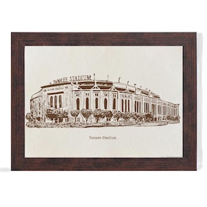Op de afbeelding: Een ingelijste prent van Yankee Stadium in sepiatinten. Het stadion is afgebeeld in een gedetailleerde lijntekening, met de woorden "Yankee Stadium" boven en onder de afbeelding. De lijst is donkerbruin.