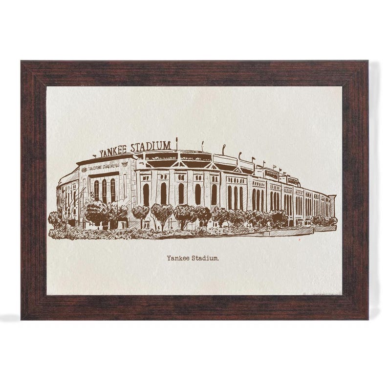 Op de afbeelding: Een ingelijste prent van Yankee Stadium in sepiatinten. Het stadion is afgebeeld in een gedetailleerde lijntekening, met de woorden "Yankee Stadium" boven en onder de afbeelding. De lijst is donkerbruin.