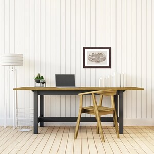 May include: A home office setup featuring a wooden desk with a black base, a laptop, and a framed architectural print. A wooden chair is positioned in front of the desk. A floor lamp and a wire basket are on the left.