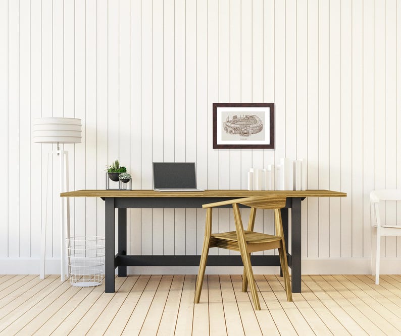 May include: A home office setup featuring a wooden desk with a black base, a laptop, and a framed architectural print. A wooden chair is positioned in front of the desk. A floor lamp and a wire basket are on the left.