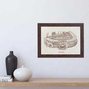 May include: A framed print of a detailed stadium illustration. The artwork, in shades of brown, depicts the stadium's architecture. The frame is dark brown, and the print is displayed on a white wall above a wooden shelf with decorative vases and books.
