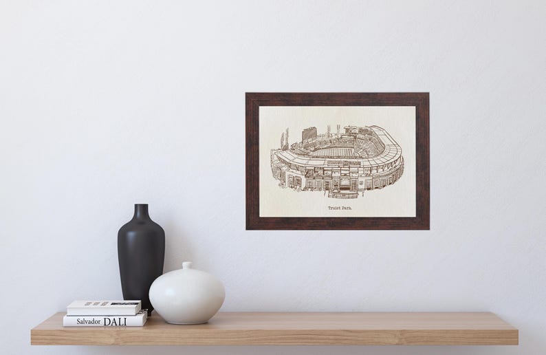 May include: A framed print of a detailed stadium illustration. The artwork, in shades of brown, depicts the stadium's architecture. The frame is dark brown, and the print is displayed on a white wall above a wooden shelf with decorative vases and books.
