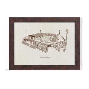 May include: A framed print featuring a detailed sepia-toned illustration of a stadium. The artwork depicts the stadium's architecture, including the stands, field, and lighting. The text "Pittbull Stadium" is printed below the image. The frame is a dark brown color.