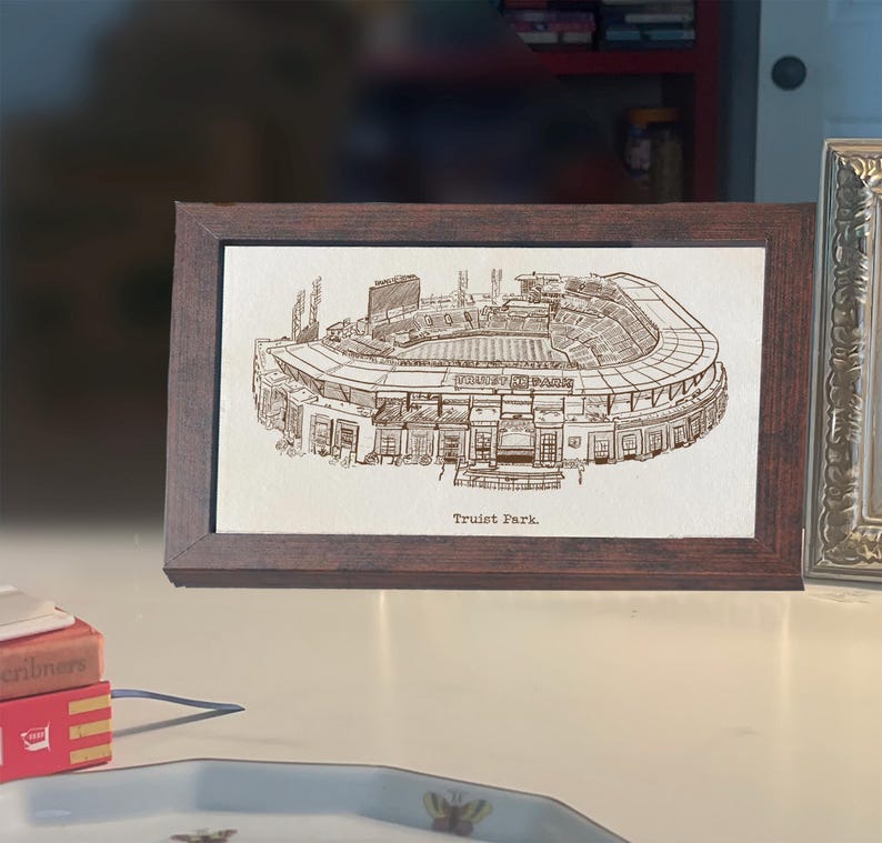 May include: A framed print of Truist Park, a detailed architectural illustration in sepia tones. The stadium is depicted with intricate lines, showcasing its structure. The frame is a dark brown, adding a classic touch to the artwork.