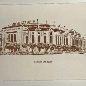Op de afbeelding: Een sepia-kleurige prent van Yankee Stadium, met gedetailleerde architectonische lijnen en de naam van het stadion bovenaan. Het kunstwerk bevat bomen en de tekst "Yankee Stadium" onderaan, tegen een getextureerde, gebroken witte achtergrond.