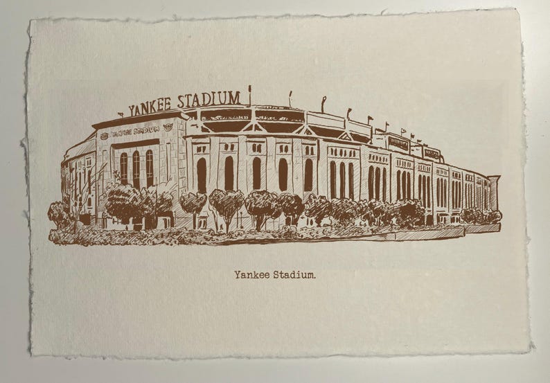 Op de afbeelding: Een sepia-kleurige prent van Yankee Stadium, met gedetailleerde architectonische lijnen en de naam van het stadion bovenaan. Het kunstwerk bevat bomen en de tekst "Yankee Stadium" onderaan, tegen een getextureerde, gebroken witte achtergrond.