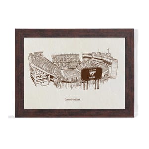 May include: A framed sepia-toned print of Lane Stadium, home of the Virginia Tech Hokies. The stadium is depicted in a detailed architectural drawing style, with the words "Lane Stadium" at the bottom. The frame is a dark brown color.