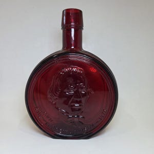 May include: A ruby red glass bottle with a round, flat body and a long neck. The front features a raised portrait of a man within a circular frame, with text around the edge. The bottle is likely vintage or antique.