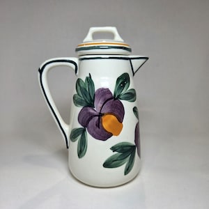 May include: White ceramic pitcher with a lid, handle, and spout. The pitcher is decorated with purple and yellow floral designs and green leaves. The rim and handle are outlined in dark blue.