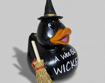 May include: A black rubber duck with a witch hat and broom. The duck has an orange beak and a pink tongue. The text "A Wee Bit WICKED" is written on the duck's body. The hat has a gold band.