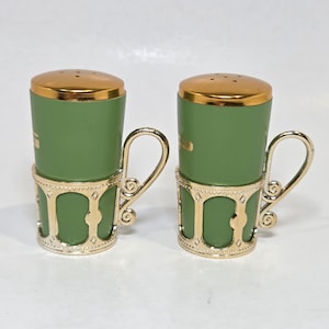 May include: A pair of green salt and pepper shakers with gold-colored lids and handles. The shakers are held in decorative gold-colored holders with a geometric design. The handles are curved and elegant, adding a touch of sophistication to the set.