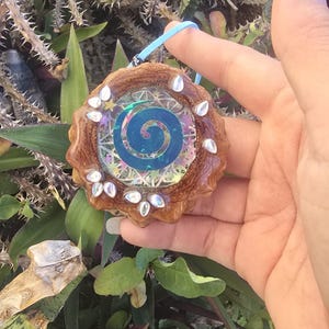 Puede incluir: Colgante artesanal con un diseño en espiral en tonos azules e iridiscentes. El colgante está engastado en un marco de madera y adornado con adornos transparentes y brillantes. Cuelga de un cordón azul claro, mostrando una estética única inspirada en la naturaleza.