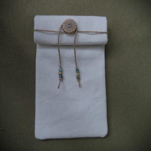 May include: A white canvas pouch with a rolled top, secured by a twine tie and a wooden button. The twine features decorative beads in shades of blue and green. The pouch is rectangular and appears to be made of a textured fabric.