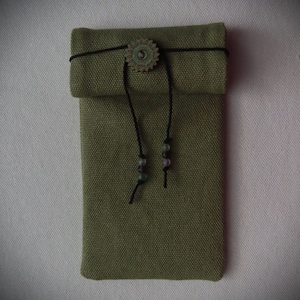 Puede incluir: Una bolsa de lona verde con un cordón de ajuste negro y un cierre de botón de metal. La bolsa tiene un acento de cuentas decorativas en el cordón.