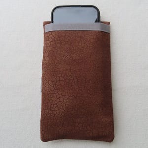 May include: A brown, textured phone sleeve with a gray elastic band at the top. The phone case is made of a material with a cracked, leather-like appearance. A black smartphone is partially visible inside the sleeve.
