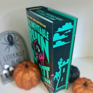 May include: A hardcover book titled "This Inevitable Ruin" by Matt Goldman. The cover is black with teal and pink accents, featuring a castle illustration. The book is displayed with pumpkins and a tombstone.