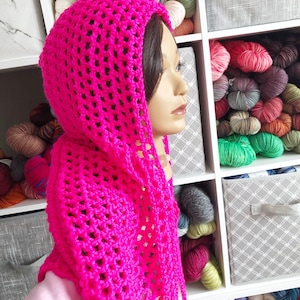 May include: A bright pink, open-weave crochet hood with long ties. The hood is displayed on a mannequin head, showcasing the texture and style of the handmade item. The background includes shelves filled with various colors of yarn.