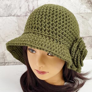 May include: Olive green crochet cloche hat with a wide brim and a decorative flower on the side. The hat is made of a textured yarn and has a classic, vintage style. The hat is shown on a mannequin head.