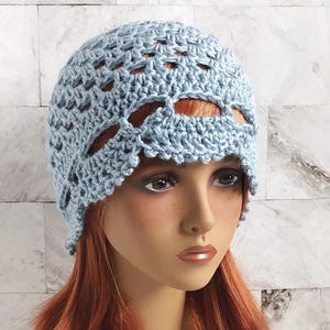May include: A light blue crochet hat with a decorative openwork design. The hat is made of a soft, textured yarn and has a scalloped edge. The hat is shown on a mannequin with red hair.