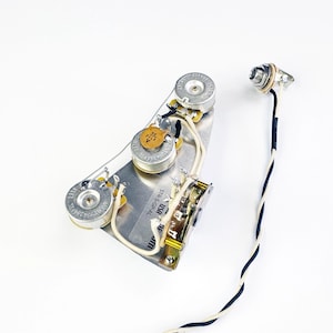 May include: A silver guitar wiring harness with three knobs, a switch, and a jack. The harness is labeled "STR 5-SG-AL 100".