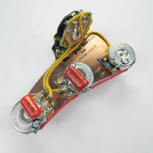 May include: A close-up of a guitar wiring harness with red and yellow wires, two red capacitors, and three potentiometers. The harness is labeled "Legend-Tone.com USA STRSSW-1V22c-CP".