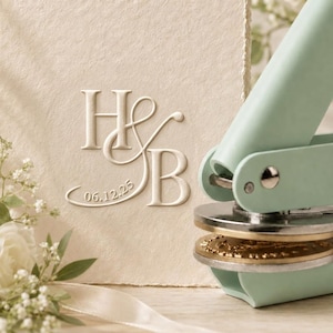 May include: A light mint green metal embosser tool positioned next to a cream-colored textured paper card. The card features a debossed monogram with the letters H and B intertwined with an ampersand and the date 06.12.25 printed below.