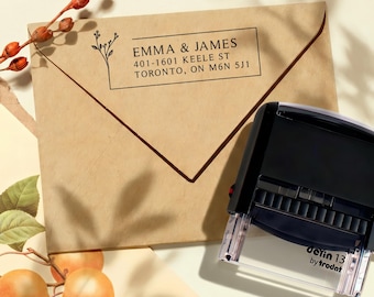 Könnte beinhalten: Ein brauner Umschlag mit dem Text "EMMA & JAMES" und Adressdetails. Ein schwarzer Stempel mit dem Text "detin 13 by trodat" befindet sich rechts. Dekorative Zweige und Früchte sind im Vordergrund.