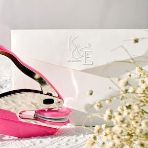 May include: A pink and silver embosser next to a white envelope with embossed initials "K&E" and the date "05.14.2025". The scene is set on a white surface with delicate white flowers in the background. The embosser is in focus, suggesting a wedding or event stationery theme.