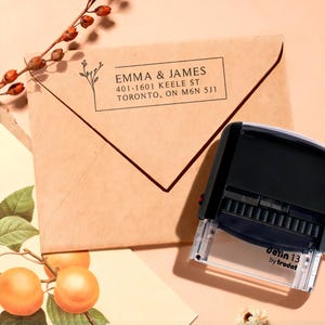 May include: A brown envelope with the text "EMMA & JAMES" and address details, along with a floral design. A black self-inking stamp with the text "Jetin 13 by trodat" is next to the envelope. Decorative sprigs and fruit are in the background.